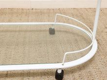 Load image into Gallery viewer, A close-up of a white metal rolling side table with a clear glass shelf and black caster wheels, resting on a woven beige rug.
