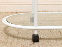 Load image into Gallery viewer, Close-up of the lower section of a white metal and glass rolling table or cart, showing a clear oval glass top, white frame, and black caster wheel on a beige woven rug.
