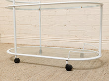 Load image into Gallery viewer, A white metal bar cart with two clear glass shelves and black caster wheels, set against a light beige brick wall and textured carpet.
