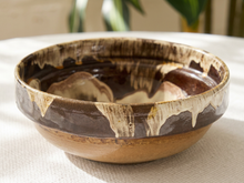 Load image into Gallery viewer, A handmade ceramic bowl with a rustic dripping glaze design, featuring a dark brown interior, a beige and brown patterned rim, and a natural tan clay base, set on a white textured tablecloth.
