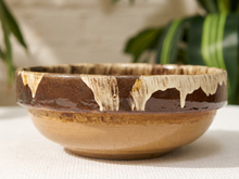 Load image into Gallery viewer, A handmade ceramic bowl with a rustic brown and beige drip glaze, displayed on a white textured surface with a blurred background of indoor plants.
