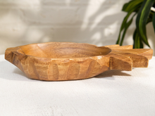 Load image into Gallery viewer, A carved wooden bowl with a handle, resting on a textured white fabric surface with a white brick wall and green plant leaves in the background.
