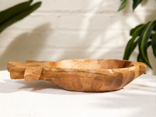Load image into Gallery viewer, A carved wooden bowl with a rustic, organic shape and visible wood grain, resting on a white textured surface with a white brick wall and green leaves in the background.
