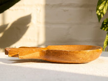 Load image into Gallery viewer, A hand-carved wooden spoon with a small integrated side dish, resting on a textured white surface with a white brick wall and green leaves in the background.
