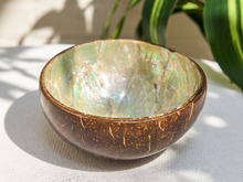 Load image into Gallery viewer, A handmade coconut shell bowl with a natural iridescent abalone shell interior, resting on a white textured fabric surface with soft leaf shadows and green leaves in the background.
