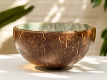 Load image into Gallery viewer, A handmade coconut shell bowl with a textured brown exterior and a green glazed interior, sitting on a white surface with soft leaf shadows.
