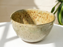 Load image into Gallery viewer, A handmade ceramic bowl with a speckled, textured glaze in earthy beige, brown, and muted green tones, sitting on a white tablecloth with soft natural shadows.
