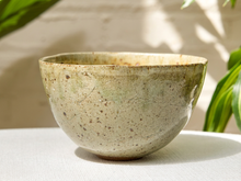 Load image into Gallery viewer, A handmade ceramic bowl with a speckled beige and light green glaze, sitting on a white textured surface with soft natural shadows and green leaves in the background.
