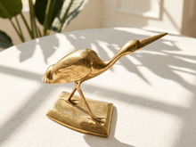 Load image into Gallery viewer, A polished brass heron figurine standing on a rectangular base, placed on a white textured tablecloth with soft sunlight and leaf shadows.

