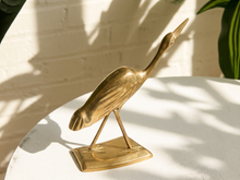 Load image into Gallery viewer, A polished brass heron figurine standing on a rectangular base, displayed on a white textured surface with warm sunlight and leaf shadows.
