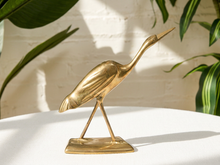 Load image into Gallery viewer, A polished brass heron figurine standing on a rectangular base, displayed on a white surface with a white brick wall and green plants in the background.
