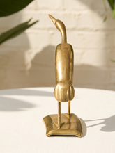 Load image into Gallery viewer, A small brass bird figurine with a long curved neck and flat feet, standing on a matching square base, photographed on a white surface against a softly lit off-white wall with blurred green leaves in the background.
