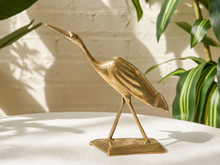 Load image into Gallery viewer, A polished brass heron figurine standing on a rectangular base, displayed on a white textured surface with a white brick wall and green houseplant leaves in the background.
