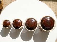 Load image into Gallery viewer, Four ceramic lidded jars in graduated sizes, arranged in a row on a white oval surface. The jars have glossy dark brown wooden lids and a light beige ceramic body, casting distinct shadows on the white surface.
