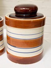 Load image into Gallery viewer, A ceramic canister with a glossy dark brown lid and brown bands, decorated with cream-colored stripes and thin blue lines, set against a white brick wall background.
