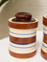 Load image into Gallery viewer, A close-up of a ceramic lidded jar with a cream body, brown bands, and blue horizontal stripes, shown on a white surface with part of a matching jar visible at the right edge.

