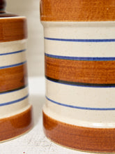 Load image into Gallery viewer, Close-up of a ceramic canister with a cream speckled body, decorated with horizontal bands of glossy brown and thin blue stripes.
