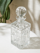 Load image into Gallery viewer, A clear cut-crystal decanter with a matching round stopper, displayed on a white textured surface with soft natural shadows and a blurred green plant in the background.
