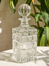 Load image into Gallery viewer, A clear cut-crystal decanter with a ribbed stopper, set on a light beige textured tablecloth with a blurred background of green houseplants and a light wall.
