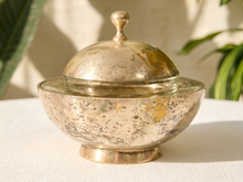 Load image into Gallery viewer, A vintage silver-toned metal lidded tureen or sugar bowl with a hammered finish, set on a white textured fabric surface with soft-focus green plants in the background.
