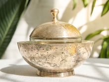 Load image into Gallery viewer, A hammered silver-toned metal lidded bowl with a glass insert, set on a white textured surface with soft leaf shadows in the background.
