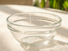 Load image into Gallery viewer, A clear glass bowl with a thick rim, resting on a textured beige fabric surface with soft natural sunlight and blurred green foliage in the background.

