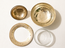 Load image into Gallery viewer, Four vintage glass and metal containers arranged on a white textured fabric background: a round gold-toned metal bowl, a larger gold-toned metal dish, a circular gold-toned metal ring with speckles, and a small clear glass bowl.
