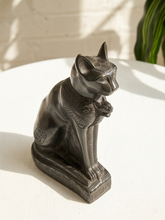 Load image into Gallery viewer, A dark metallic cat figurine with Egyptian-inspired engraved details, sitting on a white textured surface with soft shadows and a blurred green plant in the background.
