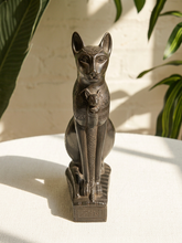 Load image into Gallery viewer, An Egyptian-style bronze cat statue of a seated cat holding a small kitten, displayed on a white surface with a white brick wall and green plants in the background.
