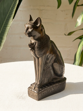 Load image into Gallery viewer, An Egyptian cat statue with a dark bronze finish, sitting on a white surface with a white brick wall and green leaves in the background.
