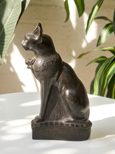 Load image into Gallery viewer, An Egyptian cat statue with hieroglyphic details, finished in a dark bronze metallic color, displayed on a white surface with leafy plants in the background.
