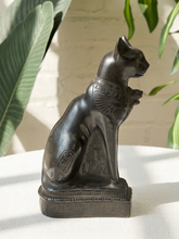 Load image into Gallery viewer, A dark bronze-finished Egyptian cat figurine, likely representing a Bastet statue, sitting on a white surface with a white brick wall and green houseplants in the background.
