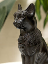 Load image into Gallery viewer, A close-up of a dark bronze-finished cat statue with carved Egyptian-style hieroglyphs on its collar, set against a softly blurred beige background with hints of green foliage.
