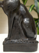 Load image into Gallery viewer, A close-up of a dark, glossy Egyptian cat statue with carved hieroglyphic details and a winged headdress, shown against a softly blurred indoor background with green leaves.
