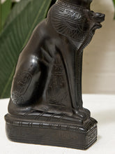 Load image into Gallery viewer, A dark carved Egyptian cat statue with hieroglyphic details, shown close-up against a soft neutral background with blurred green leaves.
