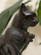 Load image into Gallery viewer, A close-up of a dark polished cat figurine with carved Egyptian-style details on its collar, shown against a softly blurred background with green leaves and a light neutral surface.
