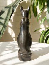 Load image into Gallery viewer, A dark bronze-finished metal sculpture of a cat viewed from the back, standing on a white textured surface with leafy green plants and a light brick wall in the background.
