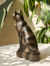 Load image into Gallery viewer, A dark bronze-finished metal sculpture of a seated cat with Egyptian-style hieroglyphic details on its collar and base, displayed on a white textured surface with leafy green plants and a light brick wall in the background.
