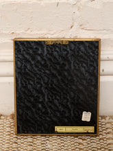 Load image into Gallery viewer, A framed black textured fabric panel, likely velvet or microfiber, with a gold-toned metal frame. The panel is displayed flat against a white brick wall on a woven beige rug, featuring a small gold label at the bottom.
