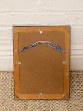 Load image into Gallery viewer, The back of a rectangular picture frame with a light brown wood border and a brown backing board, resting on a woven rug against a white brick wall.
