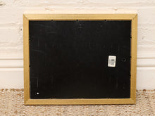 Load image into Gallery viewer, The back of a square picture frame with a gold-toned border and a black mounting board, resting on a woven rug against a white brick wall.
