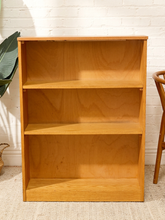Load image into Gallery viewer, A three-tier wooden bookcase with a warm honey oak finish, standing against a white brick wall on a light beige rug.
