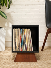 Load image into Gallery viewer, A black square record shelf on a wooden base, filled with vinyl records, set in a room with a white brick wall, beige rug, a potted plant, and a chair.
