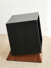 Load image into Gallery viewer, A modern black cube side table with open shelves, resting on a triangular wooden base, set on a woven rug against a white brick wall.
