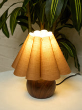 Load image into Gallery viewer, A table lamp with a pleated beige fabric shade and a round dark wood base, shown lit on a light surface with a green plant in the background.
