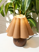 Load image into Gallery viewer, A mid-century modern table lamp with a pleated beige fabric shade and a dark wood base, styled on a white surface with a green plant in the background.
