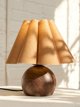 Load image into Gallery viewer, A table lamp with a pleated beige fabric shade and a polished dark brown wooden base, set against a white brick wall with soft window shadows.

