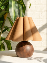 Load image into Gallery viewer, A table lamp with a pleated beige fabric shade and a round dark brown wooden base, styled on a light surface with a white brick wall and green plant leaves in the background.
