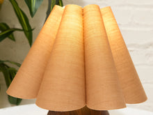 Load image into Gallery viewer, A close-up of a pleated fabric lampshade in a warm tan or light brown color, illuminated from within, set against a softly blurred background with a white brick wall and green leaves.
