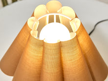 Load image into Gallery viewer, A close-up of a pleated fabric lampshade glowing warm yellow from within, with a white circular top rim and a black power cord visible on a white surface.
